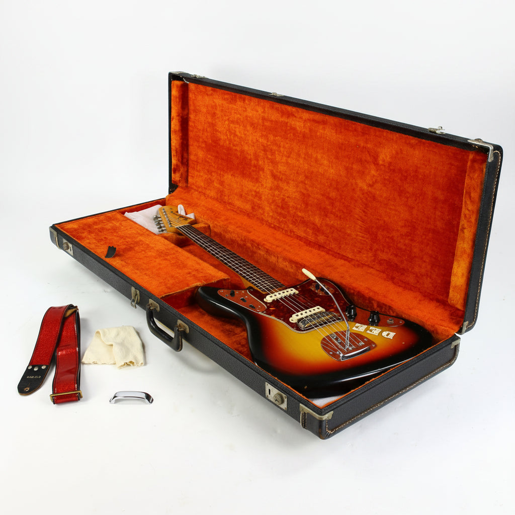 A sunburst fender jaguar electric guitar inside an open black hard case with orange velvet lining, shown on a white background with a red strap, cloth, and small accessories laid beside it.